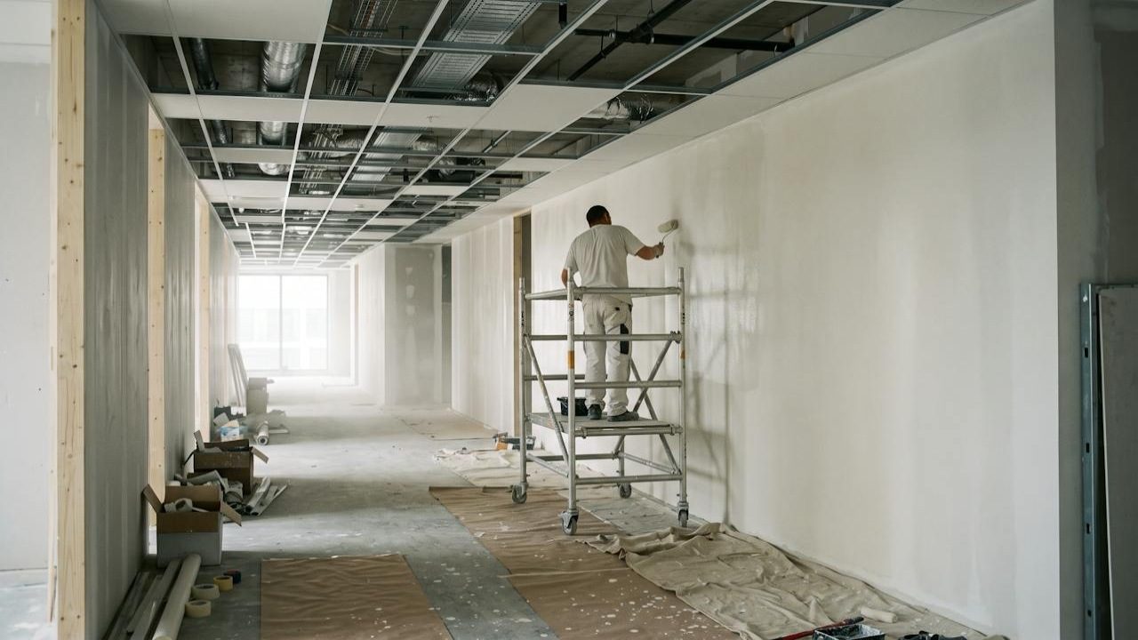 A painter on a low scaffold rolling a wall in a commercial corridor under fit-out.